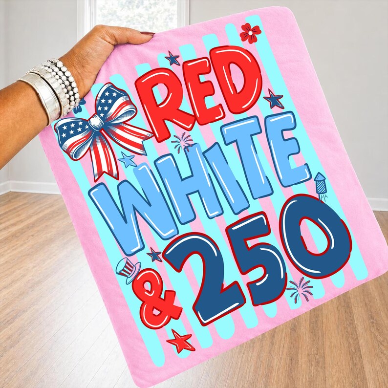 Puede incluir: Una manta rosa con rayas azules y blancas y las palabras "RED WHITE & 250" en rojo y azul. La manta tiene un lazo con un dise&ntilde;o de estrellas y rayas y otros gr&aacute;ficos patri&oacute;ticos.