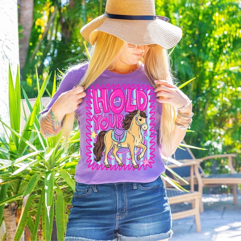 May include: A purple t-shirt with the text "HOLD YOUR HORSES" in pink and white, featuring a cartoon horse with a saddle. The shirt is paired with denim shorts and a straw hat.