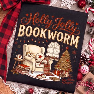 Op de afbeelding: Een zwart t-shirt met een grafische illustratie van een gezellige leeshoek met een kat op een fauteuil, een kerstboom en boeken. De tekst luidt Holly Jolly BOOKWORM en One More Chapter in decoratieve rode en cr&egrave;mekleurige letters.