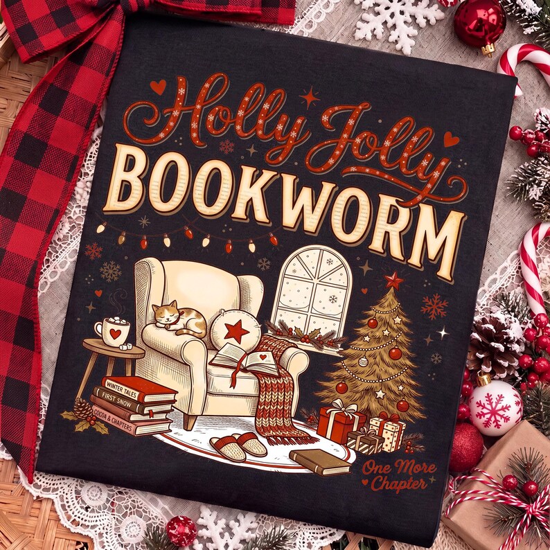 Op de afbeelding: Een zwart t-shirt met een grafische illustratie van een gezellige leeshoek met een kat op een fauteuil, een kerstboom en boeken. De tekst luidt Holly Jolly BOOKWORM en One More Chapter in decoratieve rode en cr&egrave;mekleurige letters.