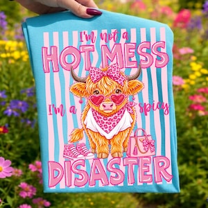 K&ouml;nnte beinhalten: Hellblaues Handtuch mit einer Cartoon-Illustration einer Highland-Kuh mit herzf&ouml;rmiger Sonnenbrille, Schleife und Halstuch. Der Text lautet: &bdquo;Ich bin kein hei&szlig;es Chaos, ich bin ein scharfes Desaster&ldquo;.