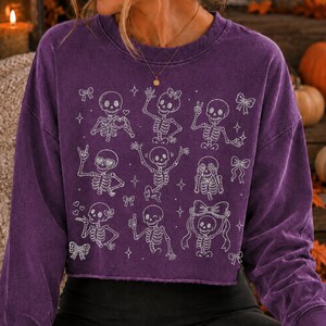 Op de afbeelding: Een paars sweatshirt met lange mouwen en ronde hals, voorzien van een witte grafische print van meerdere skeletten in diverse houdingen, zoals zwaaien, hartjeshanden en een duim omhoog, afgewisseld met kleine strikjes en sterretjes.