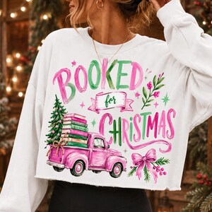 May include: A white long-sleeve sweatshirt featuring a graphic print with the text "BOOKED for CHRISTMAS". The design includes a pink pickup truck carrying a stack of books and a Christmas tree, accented with pink bows and green foliage.