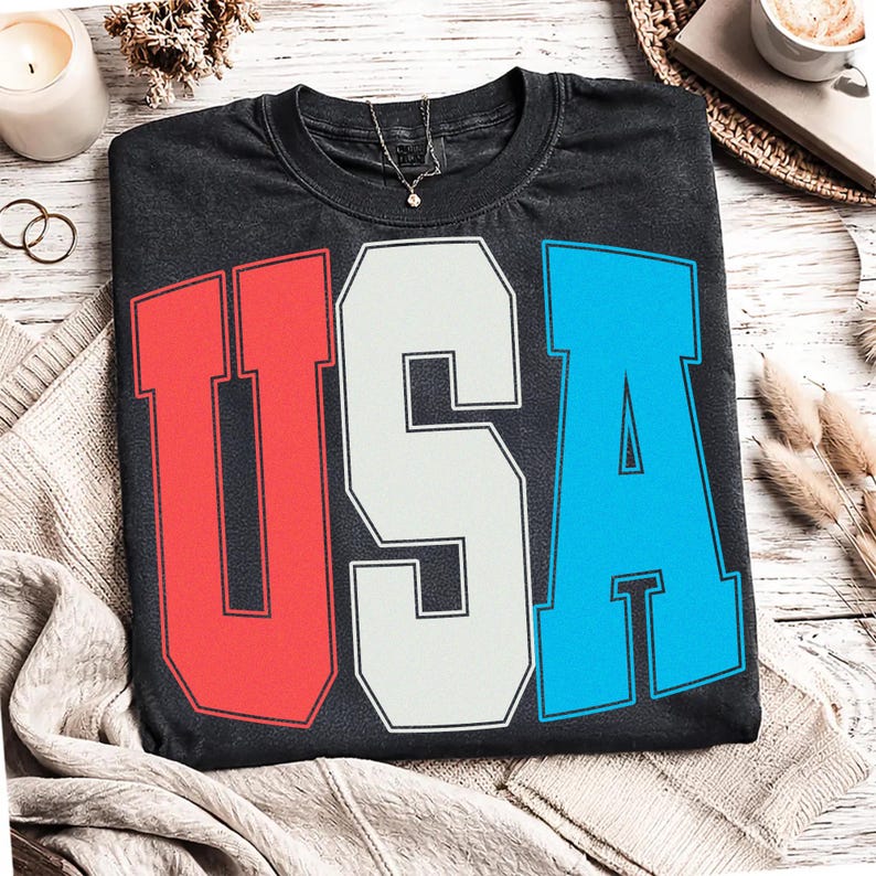 May include: A black t-shirt with the word "USA" in large, bold letters. The letters are colored red, white, and blue, with a vintage, distressed look. The shirt has a crew neck and short sleeves. A silver necklace with a small pendant is visible.
