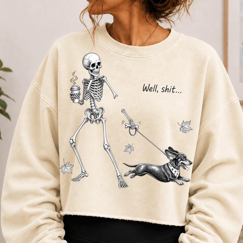 May include: A cream-colored cropped sweatshirt featuring a graphic of a skeleton holding a coffee cup and walking a dachshund on a leash. The skeleton holds a bone as a leash handle. The text "Well, shit..." and falling leaves are printed.