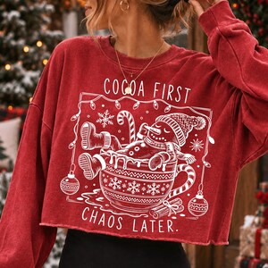Op de afbeelding: Een rood, langmouwig, kort sweatshirt met een witte afbeelding van een peperkoekmannetje dat in een mok ontspant. De tekst luidt COCOA FIRST CHAOS LATER. Het ontwerp bevat kerstverlichting, sneeuwvlokken en kerstballen in een kader.