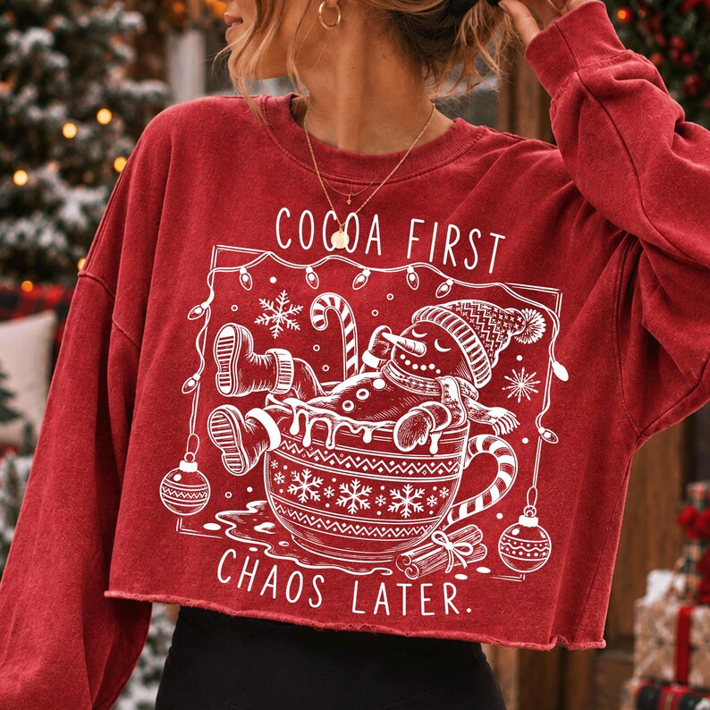 Op de afbeelding: Een rood, langmouwig, kort sweatshirt met een witte afbeelding van een peperkoekmannetje dat in een mok ontspant. De tekst luidt COCOA FIRST CHAOS LATER. Het ontwerp bevat kerstverlichting, sneeuwvlokken en kerstballen in een kader.