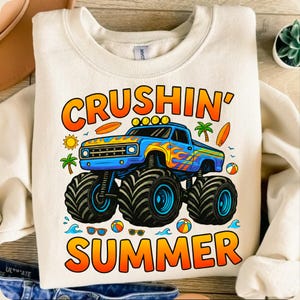 May include: Cream-colored sweatshirt with a colorful graphic of a blue monster truck with flames, palm trees, and the words "CRUSHIN' SUMMER" in orange and yellow. Includes images of a sun, surfboards, and beach balls.