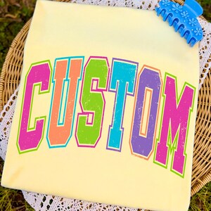 Pu&ograve; includere: Una maglietta giallo chiaro con la scritta "CUSTOM" in grandi lettere colorate in stile retr&ograve;. Le lettere sono delineate in un colore a contrasto, con un effetto sfumato. Un fermaglio blu con dettagli floreali si trova nell'angolo in alto a destra.