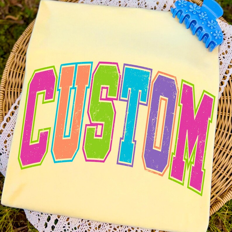 Pu&ograve; includere: Una maglietta giallo chiaro con la scritta "CUSTOM" in grandi lettere colorate in stile retr&ograve;. Le lettere sono delineate in un colore a contrasto, con un effetto sfumato. Un fermaglio blu con dettagli floreali si trova nell'angolo in alto a destra.