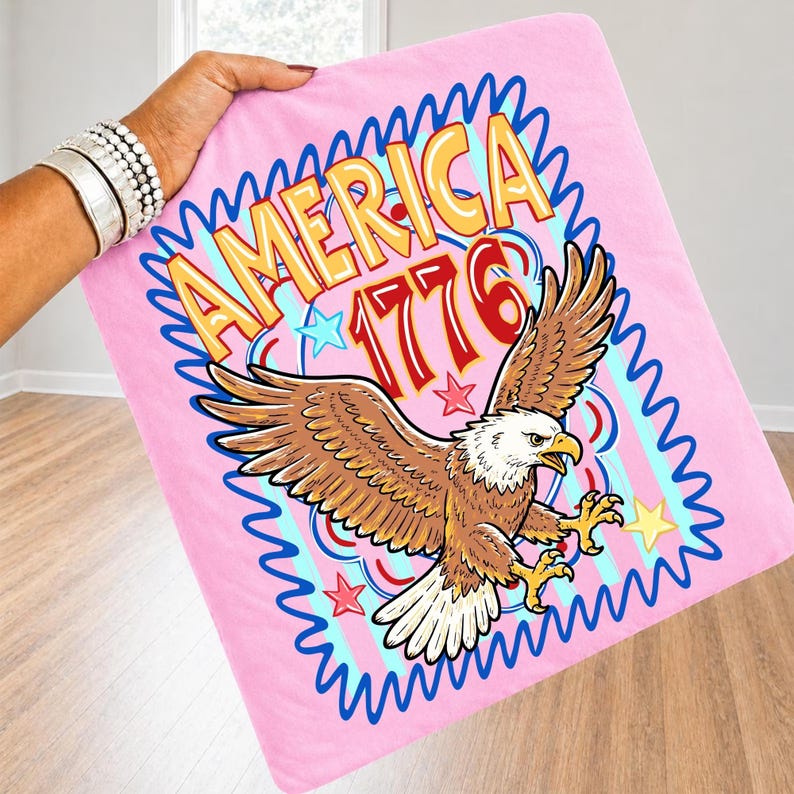 May include: Pink blanket featuring a patriotic design with the word "AMERICA" in yellow and orange, the number "1776" in red, and a brown eagle with outstretched wings. The blanket has a blue wavy border and star accents.