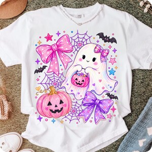 Peut inclure: Un t-shirt blanc avec une impression graphique color&eacute;e repr&eacute;sentant un fant&ocirc;me mignon tenant un seau citrouille, entour&eacute; de n&oelig;uds roses et violets, de toiles d'araign&eacute;e, de petites chauves-souris et d'&eacute;toiles. Le design utilise des tons vifs.