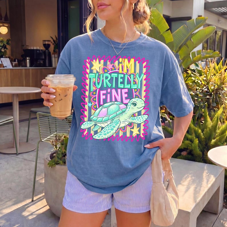 May include: A blue t-shirt with the text "Turtelly Fine" and a cartoon sea turtle graphic. The design features a pink and yellow border with stars. The shirt is paired with striped shorts.