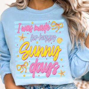 Pu&ograve; includere: Felpa azzurra con la scritta "I was made for happy sunny days" in rosa e giallo. Il design include stelle marine, fiocchi e una conchiglia. La felpa ha maniche lunghe e girocollo.
