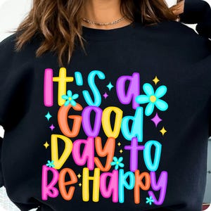 K&ouml;nnte beinhalten: Schwarzer Pullover mit einer bunten Grafik, auf der "It's a good day to be happy" mit Blumen und Sternen steht.