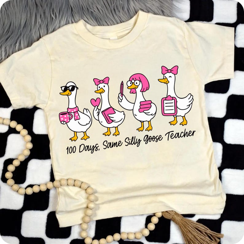 K&ouml;nnte beinhalten: Cremefarbenes T-Shirt mit einem Cartoon-Design von vier G&auml;nsen, jede mit einzigartigen Accessoires. Die G&auml;nse tragen Sonnenbrillen, Schleifen und halten Gegenst&auml;nde wie ein Herz, einen Stift und einen Klemmbrett. Der Text "100 Days. Same Silly Goose Teacher" ist darunter gedruckt.
