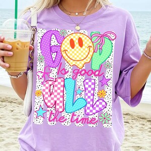 Op de afbeelding: Een lichtpaars t-shirt met een grafische print met de tekst "God is good all the time". Het ontwerp bevat een geel geblokt smileygezicht, kleurrijke stippen- en geblokte letters, roze strikjes, kleine bloemetjes en sterren op een achtergrond met zwarte stippenstrepen.