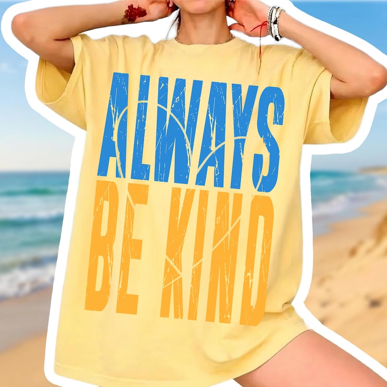 May include: Yellow t-shirt with the words "ALWAYS BE KIND" in blue and orange block letters. The shirt has short sleeves and a relaxed fit. The background shows a beach scene with blue water and sand.