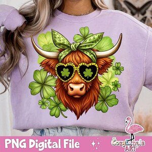 Op de afbeelding: Een paars sweatshirt met een bruine en witte Highland koe die hartvormige zonnebrillen en een groen klavertjevier-hoofdband draagt. De koe is omringd door groene klaverblaadjes.