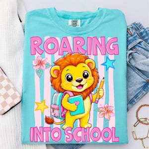 May include: A light blue t-shirt featuring a graphic of a cartoon lion with a brown mane, wearing a pink backpack, and holding a pencil and a book. The text ROARING INTO SCHOOL is printed in pink. The background has vertical pink and blue stripes.