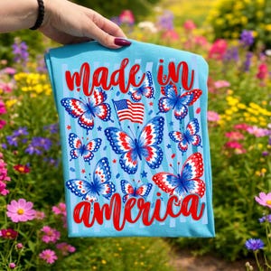 May include: A light blue t-shirt featuring a repeating pattern of butterflies with red, white, and blue wings. The design includes a small American flag and the red cursive text "made in america" printed at the top and bottom of the graphic.