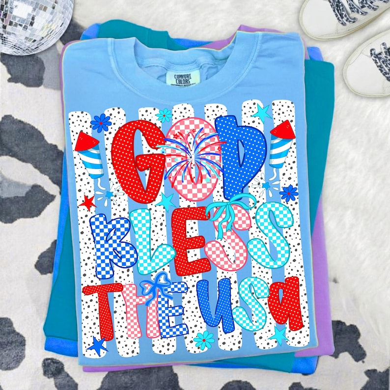 May include: A light blue t-shirt featuring the text "GOD BLESS THE USA" in a mix of red, white, and blue patterns including polka dots and checkers. The design includes firework graphics, small stars, and flowers over vertical black dots.
