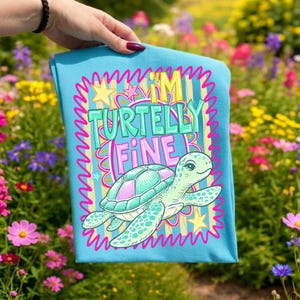 May include: A light blue t-shirt featuring a cartoon turtle with a teal and pink shell. The shirt has the text "I'M TURTELLY FINE" in a colorful, playful font. The design is framed by a wavy pink border and yellow stars.