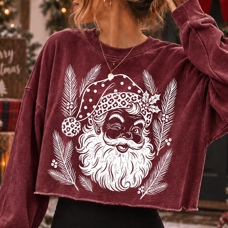 Puede incluir: Una sudadera corta de manga larga color granate con un estampado gr&aacute;fico blanco de un Santa Claus gui&ntilde;ando un ojo y usando un gorro con estampado de leopardo. El dise&ntilde;o est&aacute; rodeado de ramas de pino blancas y peque&ntilde;as bayas.