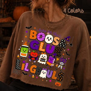 K&ouml;nnte beinhalten: Ein braunes, lang&auml;rmeliges, bauchfreies Sweatshirt mit einer bunten Halloween-Grafik und dem Text "here for the BOOS & GLUE with my LIL GHOULS". Das Design enth&auml;lt Cartoon-Illustrationen eines Geistes, einer Klebeflasche, eines Apfels, eines Buches und einer Katze.