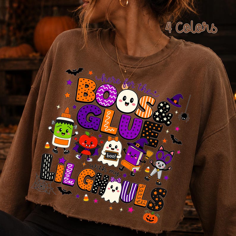 K&ouml;nnte beinhalten: Ein braunes, lang&auml;rmeliges, bauchfreies Sweatshirt mit einer bunten Halloween-Grafik und dem Text "here for the BOOS & GLUE with my LIL GHOULS". Das Design enth&auml;lt Cartoon-Illustrationen eines Geistes, einer Klebeflasche, eines Apfels, eines Buches und einer Katze.