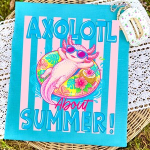 Peut inclure: Une serviette turquoise avec des rayures verticales roses et les mots "AXOLOTL ABOUT SUMMER!" en bleu et rose. Le motif repr&eacute;sente un axolotl de dessin anim&eacute; portant des lunettes de soleil et flottant sur une bou&eacute;e avec une boisson tropicale.