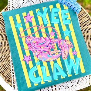 May include: Teal pillow with yellow vertical stripes and the words "YEE CLAW" in turquoise block letters. A pink and blue lobster wearing a cowboy hat is in the center, surrounded by pink and turquoise stars.