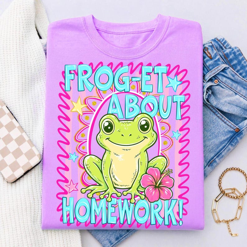 K&ouml;nnte beinhalten: Ein lilafarbenes T-Shirt mit einem Frosch-Cartoon und dem Text "FROGET ABOUT HOMEWORK!". Der Frosch ist gr&uuml;n und gelb und h&auml;lt eine rosa Blume. Der Text ist t&uuml;rkis, mit einem rosa-gelben Blumenrand.
