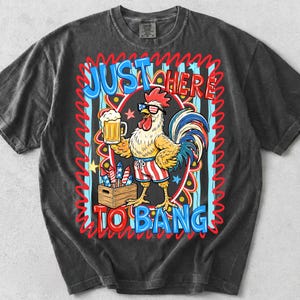 May include: Dark gray t-shirt featuring a cartoon rooster wearing sunglasses and patriotic shorts, holding a beer and fireworks. The shirt has the text "JUST HERE TO BANG" in a colorful design.