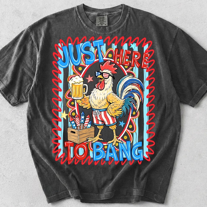 May include: Dark gray t-shirt featuring a cartoon rooster wearing sunglasses and patriotic shorts, holding a beer and fireworks. The shirt has the text "JUST HERE TO BANG" in a colorful design.