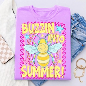K&ouml;nnte beinhalten: Ein helllila T-Shirt mit einem Cartoon-Bienen-Motiv und dem Text "BUZZIN INTO SUMMER!" in Gelb. Die Biene ist gelb und schwarz mit blauen Fl&uuml;geln, umgeben von Sternen und einem rosa Blumendesign. Das T-Shirt ist gefaltet.