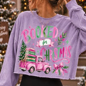 May include: A light purple long-sleeve sweatshirt featuring a graphic print with the text BOOKED for CHRISTMAS. The design includes a pink pickup truck carrying a stack of books and a Christmas tree, accented with green foliage and a pink bow.