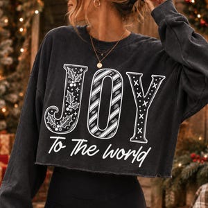 May include: A charcoal gray long-sleeve cropped sweatshirt featuring the white text JOY with holly leaves in the J, diagonal stripes in the O, and X patterns in the Y. Below the word JOY, the text To The world is printed in white script.