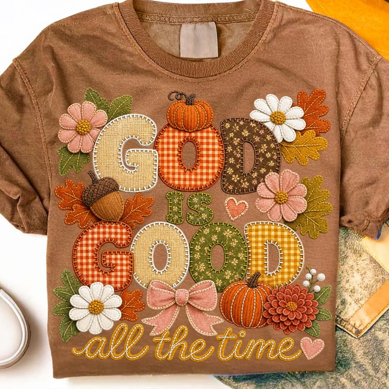 Pu&ograve; includere: Una t-shirt marrone a maniche corte con una grafica che riporta il testo "GOD IS GOOD all the time". Il design include vari motivi come il vichy e fiori, insieme a illustrazioni di zucche, foglie, una ghianda e un fiocco.
