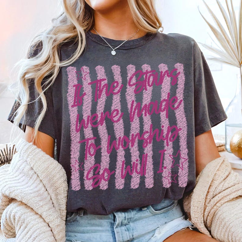 May include: Dark gray t-shirt with the text "If The Stars Were Made To Worship So Will I" in pink. The shirt has a crew neck and short sleeves. The text is in a vertical striped design.