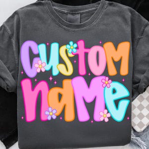 May include: Dark gray sweatshirt with the words "CUSTOM NAME" in colorful bubble letters. The letters are outlined in various colors, including purple, blue, pink, and orange, and adorned with flower graphics. The design is centered on the front of the shirt.