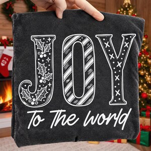 May include: A black t-shirt featuring the white text JOY in a large, stylized font. The letter J is decorated with holly leaves and berries, the O has diagonal stripes, and the Y has an X pattern. Below, the text To The world is printed.