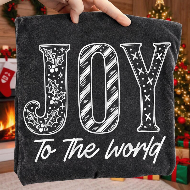 May include: A black t-shirt featuring the white text JOY in a large, stylized font. The letter J is decorated with holly leaves and berries, the O has diagonal stripes, and the Y has an X pattern. Below, the text To The world is printed.