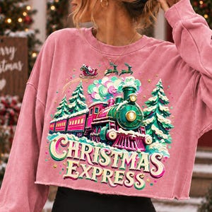 May include: A dusty pink long-sleeve cropped sweatshirt featuring a graphic print of a green and red steam train travelling through a snowy forest. Above the train, Santa Claus flies in a sleigh with reindeer. The text reads CHRISTMAS EXPRESS.