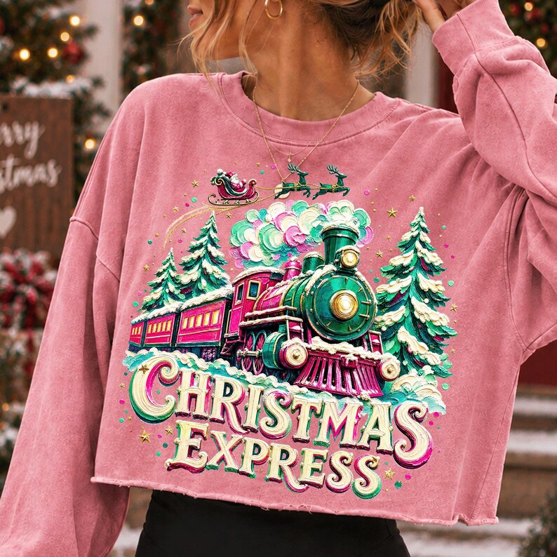 May include: A dusty pink long-sleeve cropped sweatshirt featuring a graphic print of a green and red steam train travelling through a snowy forest. Above the train, Santa Claus flies in a sleigh with reindeer. The text reads CHRISTMAS EXPRESS.