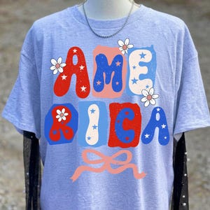 May include: A pale blue t-shirt featuring the word "AMERICA" in red, blue, and white block letters. The letters are decorated with white stars and flowers, and a pink bow is at the bottom. The shirt is a casual style.