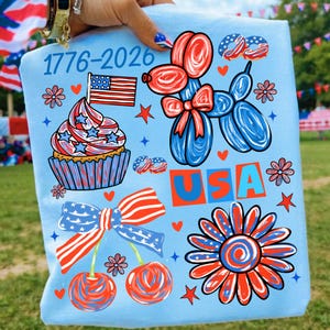 Pode incluir: Camiseta azul clara com gr&aacute;ficos patri&oacute;ticos. Apresenta um cupcake com uma bandeira, um c&atilde;o bal&atilde;o, cerejas e uma flor, todos em vermelho, branco e azul. O texto "1776-2026" e "USA" tamb&eacute;m s&atilde;o exibidos.