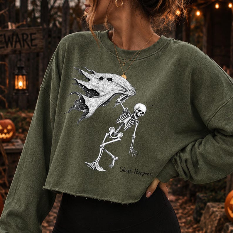 K&ouml;nnte beinhalten: Ein olivgr&uuml;nes, lang&auml;rmeliges, bauchfreies Sweatshirt mit einem Grafikdruck eines Skeletts, das ein wei&szlig;es Laken mit himmlischem Muster h&auml;lt. Der Text "Sheet Happens..." ist in schwarzer Schreibschrift unter dem Skelett abgedruckt.