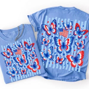 Puede incluir: Una camiseta azul claro con un estampado gr&aacute;fico de varias mariposas en patrones de color rojo, blanco y azul, intercaladas con peque&ntilde;as estrellas y una representaci&oacute;n de la bandera estadounidense. El dise&ntilde;o se sit&uacute;a sobre rayas verticales azul claro.