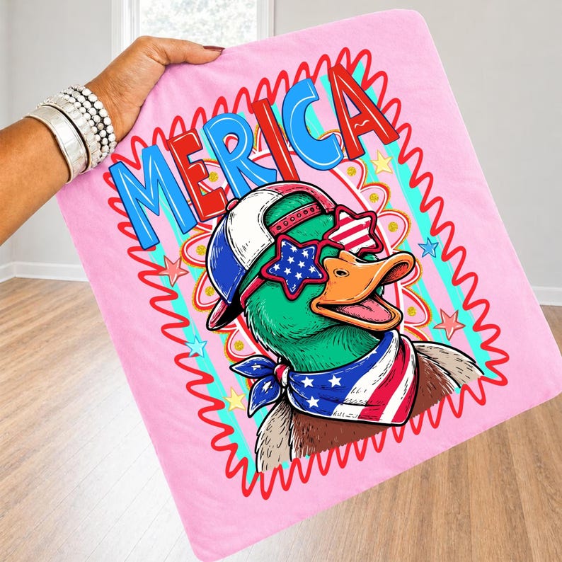 May include: Pink blanket with a cartoon duck wearing sunglasses, a hat, and a bandana, all in patriotic colors. The word "MERICA" is displayed in a red and blue font, surrounded by a wavy border.